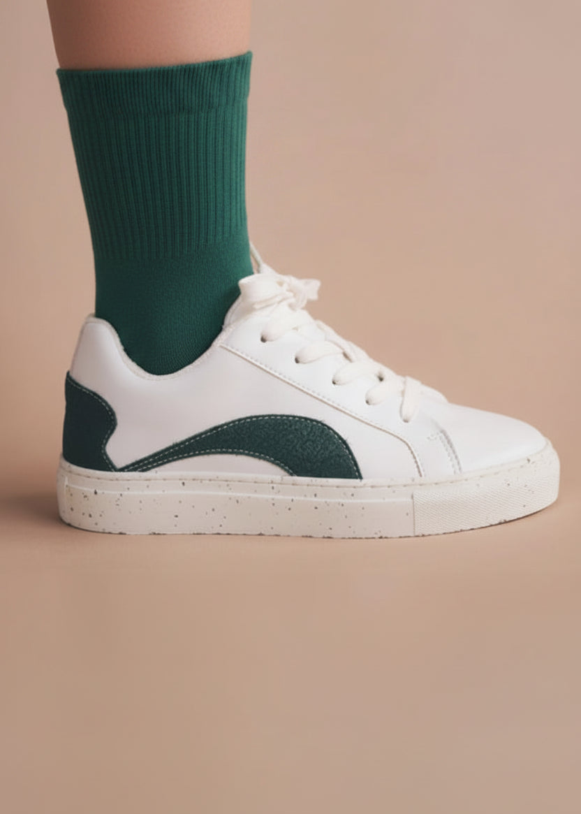 A person's lower leg wearing a lime green ribbed crew sock with and a white low-top sneaker with a dark green heel accent, set against a solid dusty pink background.