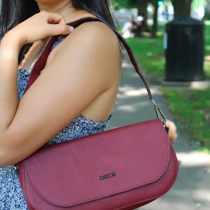 Person holding a maroon handbag with a blurred park background. plant based garnet colored leather bad made from apple peels.  Classic shoulder bag with fold-over design