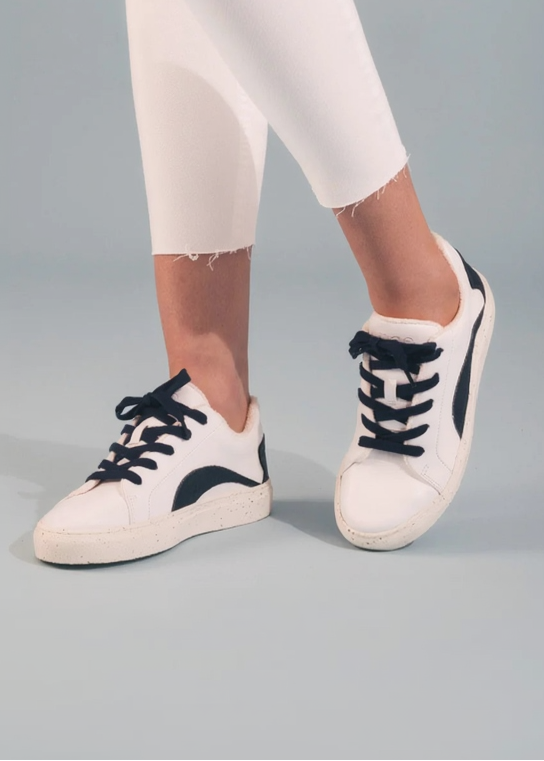 White low-top sneakers with black laces and black side accents, shown on a model wearing white cropped jeans against a light gray studio background.